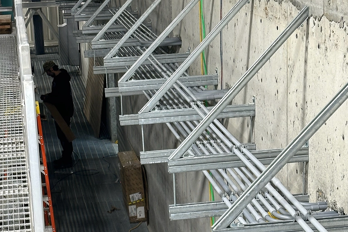 Electrician working along a wall with mounted conduit supports and organized metal framing for cable management.